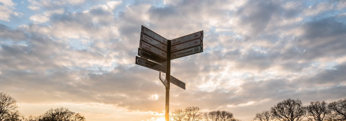 Ein Wegweiser mit mehreren Richtungsschildern vor einem Abendhimmel mit Wolken und tief stehender Sonne
