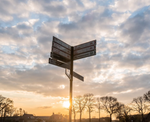 Ein Wegweiser mit mehreren Richtungsschildern vor einem Abendhimmel mit Wolken und tief stehender Sonne